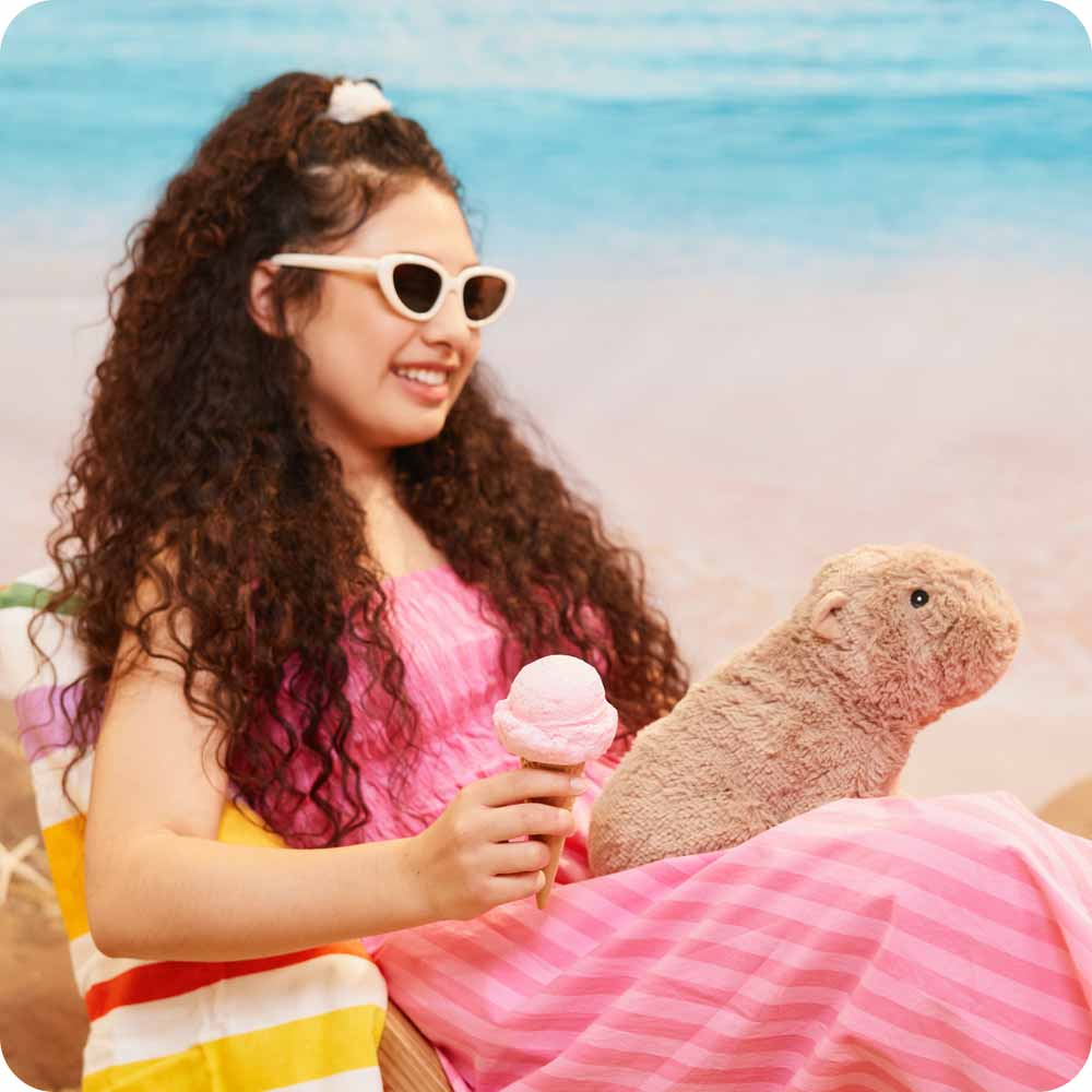 Image 104: Young teenage girl holding an icecream while sitting on the beach with a Capybara Warmies in her lap
